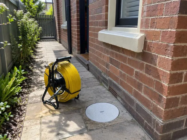 Capped drain inspection opening beside a coiled camera reel on a paved side path by a brick house in Randwick