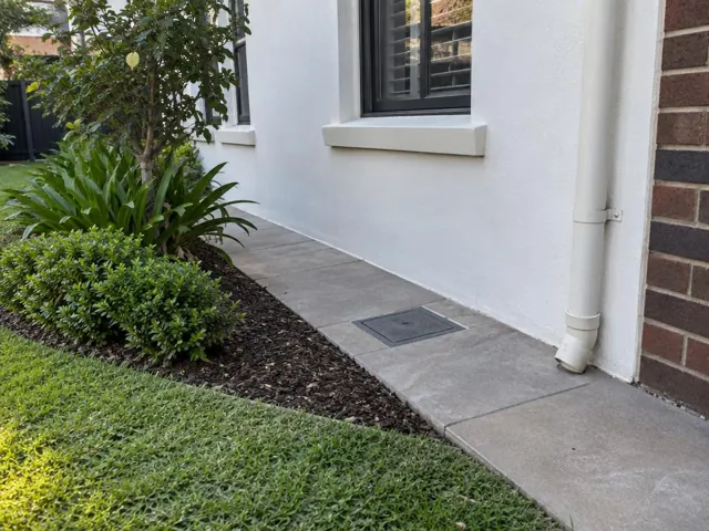 Flush access cover set in a paved side path beside a white house wall and garden bed in Kensington